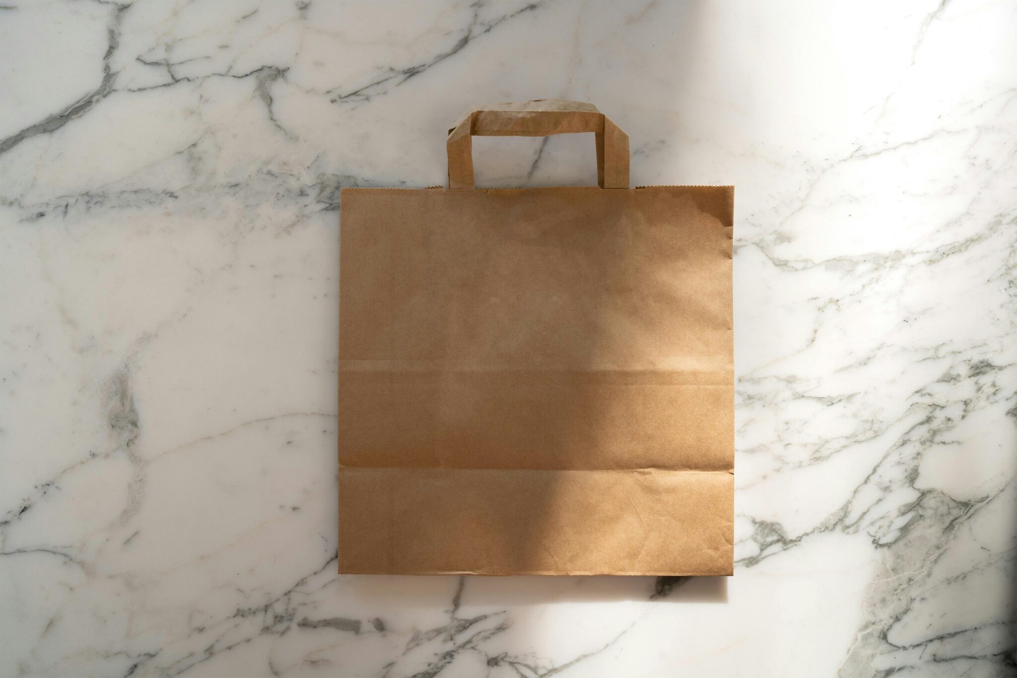 A brown paper bag with handles on a white marble surface, illuminated by natural light.